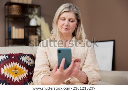 Relaxed mature old 60s woman middle aged female holding smartphone using mobile app, texting message, search ecommerce offers on cell phone technology device sitting on couch at home.
