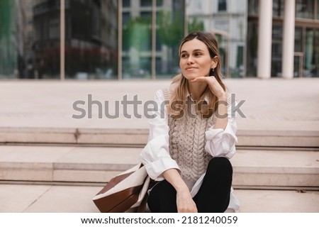 Attractive blonde woman sitting on stairs near office building and look happy. Blonde girl smiling and look at side. Amazed woman wear beige knit vest, white shirt.