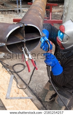 A professional welder in a protective suit and mask produces the connection of pipes with the help of a welding machine on the construction site.