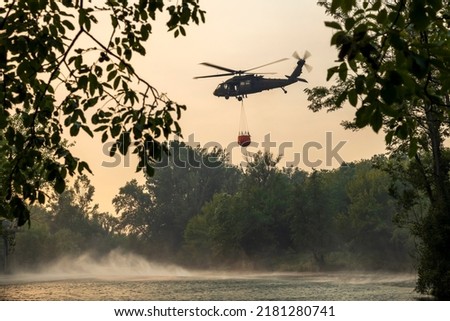 Military Chopper Helps in Firefighting Loading Water from a River into a Firefighter Bucket - Sky full of Extensive Blaze Smoke in Mid Afternoon