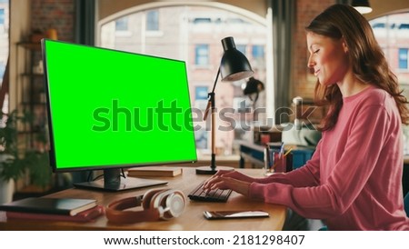 Young Beautiful Adult Woman Working from Home on Desktop Computer with Green Screen Mock Up Display. Creative Female Checking and Writing Emails. Loft Apartment with Urban City View from Big Window.