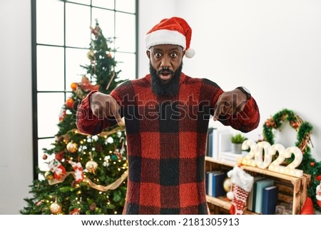 African american man wearing santa claus hat standing by christmas tree pointing down with fingers showing advertisement, surprised face and open mouth 