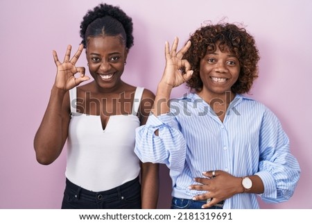 Two african women standing over pink background smiling positive doing ok sign with hand and fingers. successful expression. 
