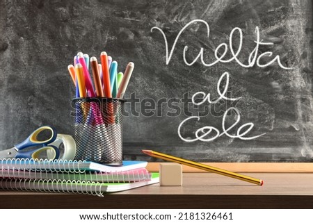 School supplies on wooden table and blackboard in the background and message vuelta al cole. Front view. Horizontal composition. Royalty-Free Stock Photo #2181326461
