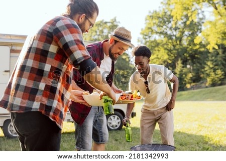A group of friends spend time together in nature. The middle-aged men prepare a barbecue near the RV. A bearded boy carries snacks for the barbecue.