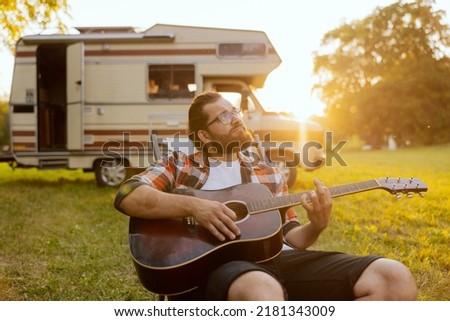A handsome bearded man plays guitar not far from his beloved RV.