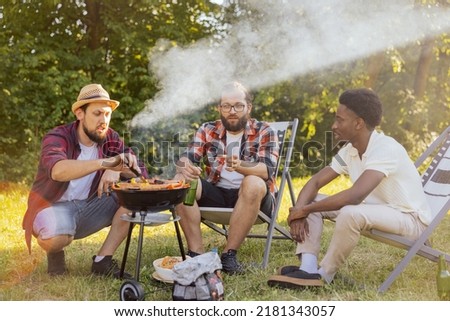 A group of friends spend time together in nature. The middle-aged men prepare a barbecue near the RV. Bearded boy checks the grill