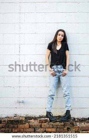 A teenage girl with long hair wearing black, denim and combat boots leaning against an old cinder block wall and looking down with attitude.