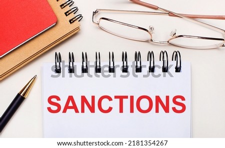 On a light background - glasses in gold frames, a pen, brown and red notepads and a white notebook with the text SANCTIONS. Business concept