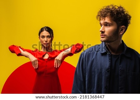 trendy curly man near blurred woman in dress holding round shape glass isolated on yellow