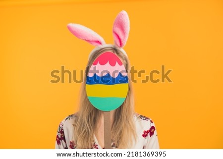 Unrecognizable caucasian person with long blond hair and white floral blouse has white bunny ears on their head and covers face with colourful cardboard Easter egg. High quality photo