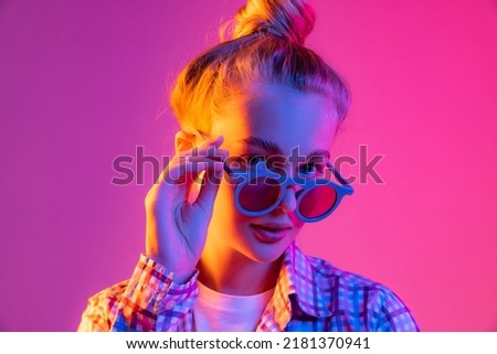 Party. Pretty young girl, student in cool eyeglasses wearing plaid shirt isolated on magenta color background in neon light. Concept of beauty, art, fashion, youth, sales and ad, education, studying. Royalty-Free Stock Photo #2181370941