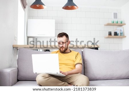 Bearded man wearing yellow shirt working or shopping online using laptop at home sitting on a cozy sofa. Online shopping, home office, remote work or education concept. Selective focus.