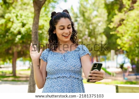 Young brunette girl smiling happy wearing summer dress on city park, outdoors holding phone looking at screen waving hand video calling distance friend online in mobile app using phone video chat app.