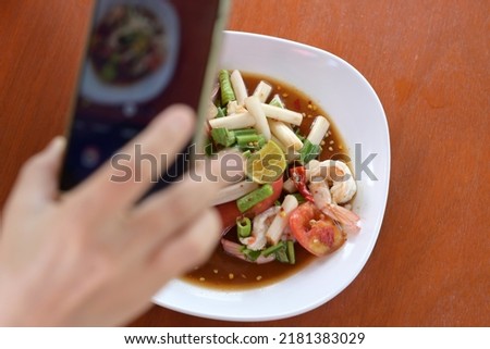 Lotus root Stem Thai Salad with Shrimp on the table. Blurred image of a woman's hand holding Mobile phone taking pictures of Lotus Stem Thai Salad on the table. Focus on food. Similar to papaya salad,