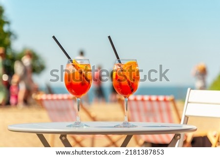 Two glasses of orange spritz aperol drink cocktail on table outdoors with sea and trees view blurred people background. Royalty-Free Stock Photo #2181387853