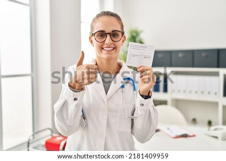 Young doctor woman holding covid certificate smiling happy and positive, thumb up doing excellent and approval sign 