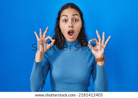 Young brazilian woman standing over blue isolated background looking surprised and shocked doing ok approval symbol with fingers. crazy expression 