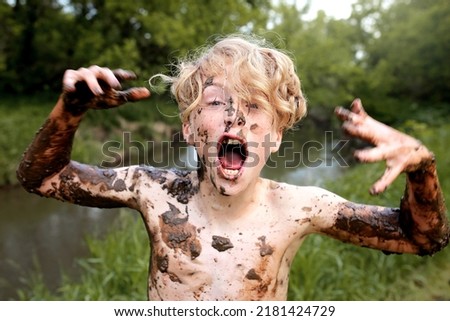 A wild kid is happily yelling while covered in mud after swimming in the river. Royalty-Free Stock Photo #2181424729