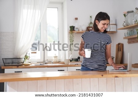 A woman in the kitchen at the computer with a smile. A woman types on a computer, works from home or communicates on social networks. A woman drinks tea and holds a cup in her hand. High quality photo