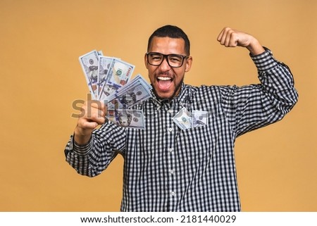Happy winner! Young rich african american man in casual holding money dollar bills with surprise isolated over beige background.