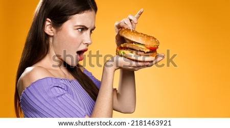 Young woman looks inside burger with surprised face. Girl is shocked after seeing cheeseburger ingredients, orange background