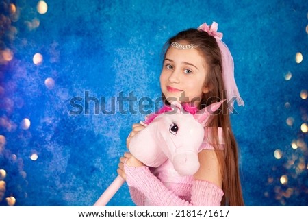 Portrait of a cute positive happy teenage girl in a pink ball gown playing with a unicorn toy. Photo on a blue background with bokeh and confetti.