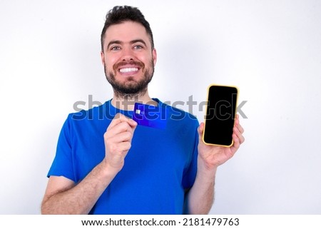 Photo of adorable Young caucasian man wearing blue T-shirt over white background holding credit card and Smartphone. Reserved for online purchases