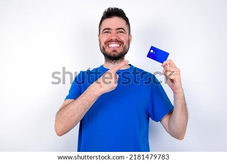 Photo portrait of Young caucasian man wearing blue T-shirt over white background doing purchase with pointing finger credit bank card