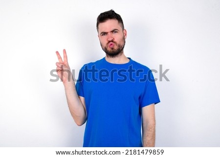 Young caucasian man wearing blue T-shirt over white background makes peace gesture keeps lips folded shows v sign. Body language concept