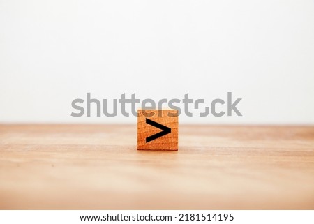 Greater. The inequality sign. Comparison operator. Written on a wooden block. Black letters. Wooden table background.