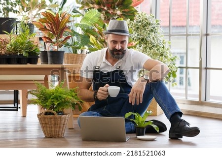 A bearded man wearing hat sitting and drinking coffee and using laptop notebook computer to sell plant in home indoor garden. Online marketing and live stream seller concept.