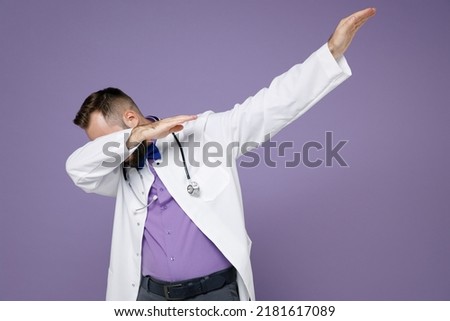 Young bearded doctor man in medical gown doing dab hip hop dance hands gesture, youth sign hiding and covering face isolated on violet background studio. Healthcare personnel health medicine concept