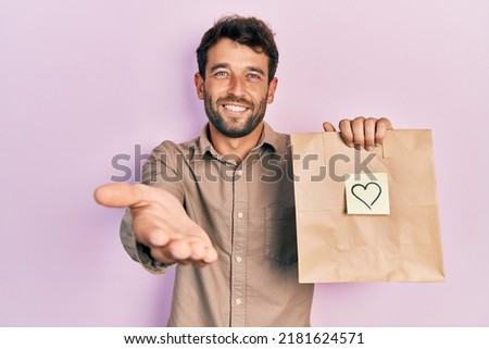 Handsome man with beard holding delivery paper bag with heart reminder smiling cheerful offering palm hand giving assistance and acceptance. 