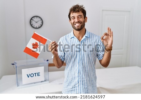 Young handsome man at political campaign election holding canada flag doing ok sign with fingers, smiling friendly gesturing excellent symbol 