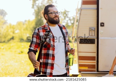 A bearded middle-aged boy emerges from an RV. The student boy is holding a bottle of his favorite beer and a guitar