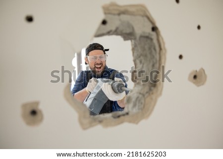 Crazy energetic man vandalizes apartment with impact hammer forges wall punches a hole through concrete, house construction site. Royalty-Free Stock Photo #2181625203
