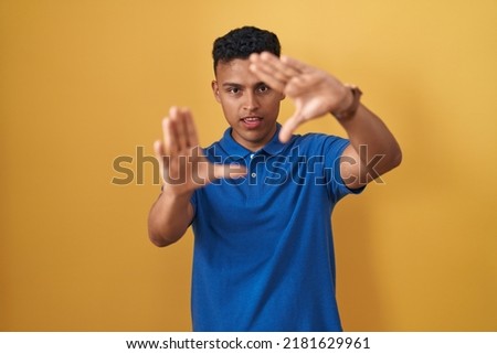 Young hispanic man standing over yellow background doing frame using hands palms and fingers, camera perspective 