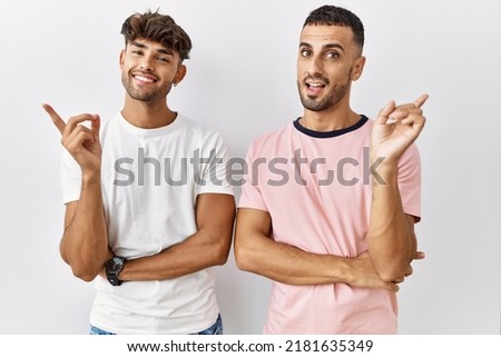 Young gay couple standing over isolated background with a big smile on face, pointing with hand finger to the side looking at the camera. 
