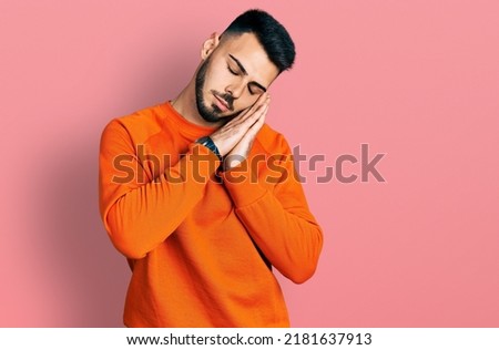 Young hispanic man with beard wearing casual orange sweater sleeping tired dreaming and posing with hands together while smiling with closed eyes. 