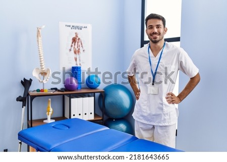 Young hispanic man wearing physiotherapist uniform standing at rehab clinic Royalty-Free Stock Photo #2181643665