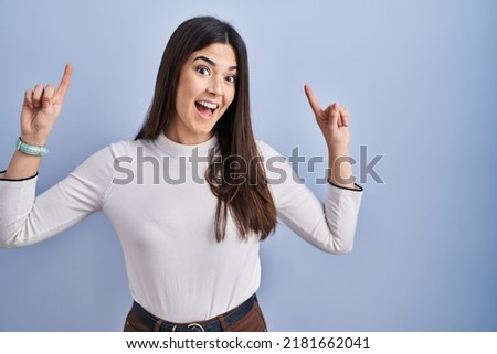 Young brunette woman standing over blue background smiling amazed and surprised and pointing up with fingers and raised arms. 