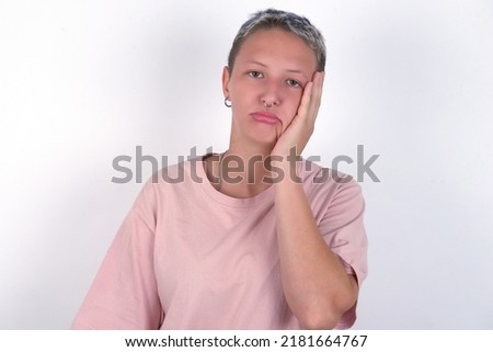 Sad lonely young woman with short hair wearing pink t-shirt over white background touches cheek with hand bites lower lip and gazes with displeasure. Bad emotions