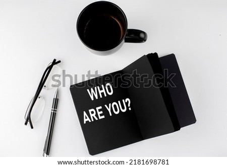 WHO ARE YOU written text in a small black notebook with coffee , pen and glasess on white background. Black-white style