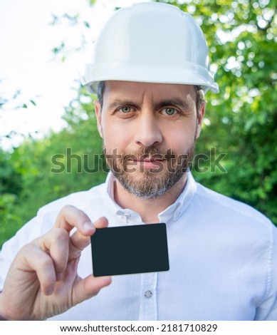 Man architect in hardhat showing blank contact card outdoors, copy space