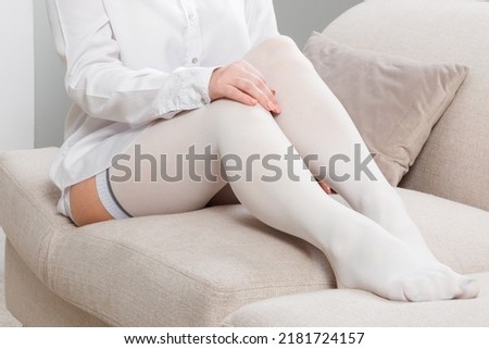 White hosiery. Beautiful long female legs in stockings. Girl putting on stockings at home in a white room. Varicose veins prevention. Woman body in underwear.