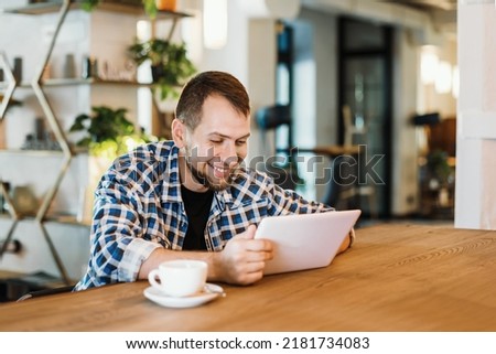 A young man in a plaid shirt drinks coffee and uses a tablet and a phone. Hipster with gadgets