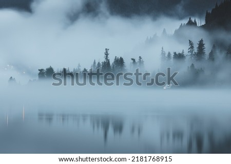 Moody, atmospheric evening fog over Kootenay Lake in Nelson, BC, Canada, with a remote, secluded cabin along the waterfront. Royalty-Free Stock Photo #2181768915