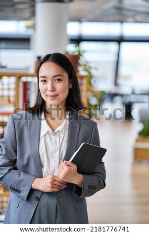Smiling young Asian business woman wearing suit holding digital tablet standing in office. Professional executive manager or teacher using corporate technology looking at camera. Vertical portrait Royalty-Free Stock Photo #2181776741