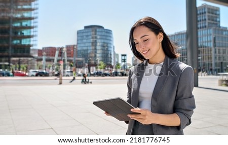 Young happy Asian professional woman wearing suit holding digital tablet standing in big city on busy street, smiling lady using smart business software for online work on pad computer outside. Royalty-Free Stock Photo #2181776745